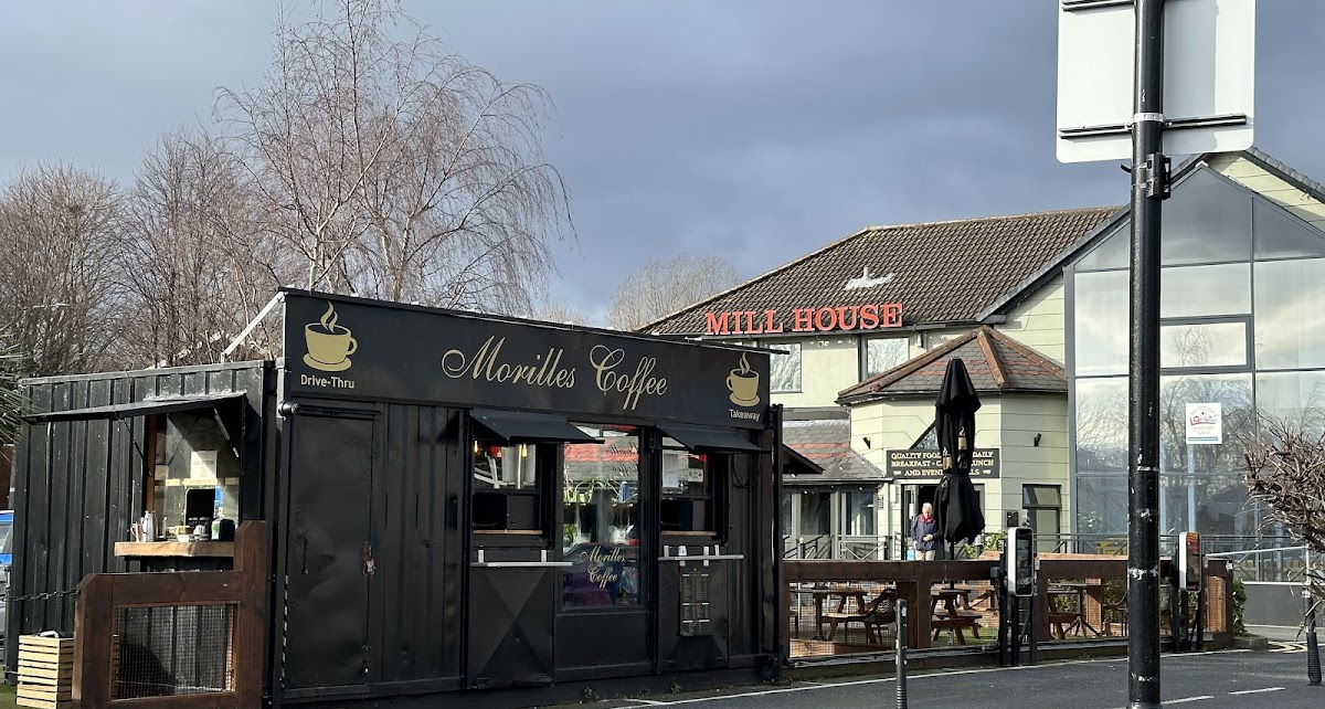 Drive-thru coffee stand near Mill House restaurant, situated in a parking area with a cloudy sky backdrop.