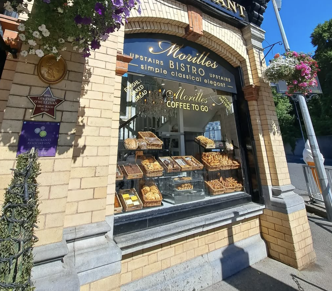 Bistro storefront with pastries on display, offering coffee to go; brick exterior and hanging flowers enhance charm.